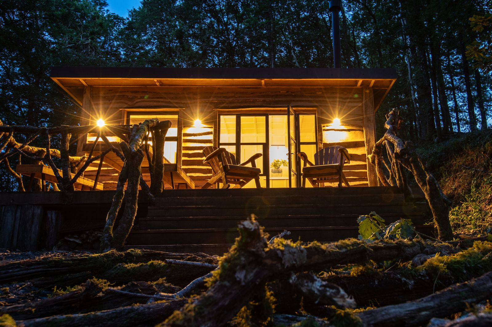 Illuminated boutique woodland cabin at dusk with glowing warm lighting and timber terrace